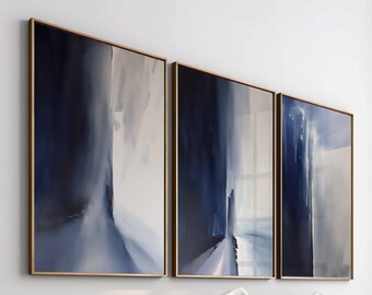 May include: Three abstract paintings in a modern bedroom setting. The paintings are framed in gold and feature shades of blue and white. The paintings are hung above a bed with a white duvet and brown throw blanket.