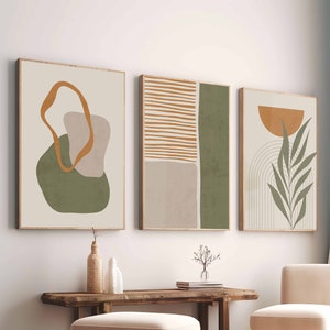 Könnte beinhalten: Drei abstrakte Kunstdrucke mit minimalistischem Design, die grüne, braune und orangefarbene Formen und Linien zeigen. Die Drucke sind in Holz gerahmt und an einer weißen Wand über einer Holzbank und einem weißen Stuhl aufgehängt.