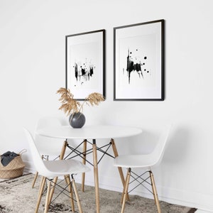 May include: Two black framed abstract art prints with white backgrounds and black ink splatter designs hang on a white wall above a white round table with four white chairs.