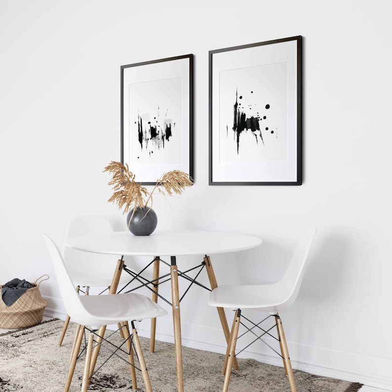 May include: Two black framed abstract art prints with white backgrounds and black ink splatter designs hang on a white wall above a white round table with four white chairs.