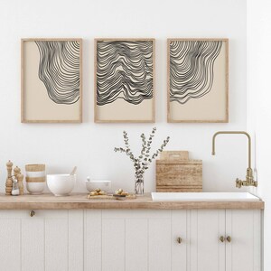 May include: Three abstract line art prints in wooden frames, hung on a white wall above a kitchen counter with a white sink and a gold faucet.