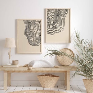 May include: Two framed black and white line art prints with a beige background. The prints are hanging on a white wall above a wooden bench with a white pillow. A potted plant with green leaves is on the right side of the bench.