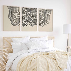 May include: Three abstract line art prints in black and white on a beige background. The prints are framed and hung on a white wall above a bed with white bedding and a cream throw blanket.