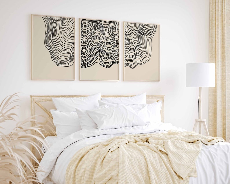 May include: Three abstract line art prints in black and white on a beige background. The prints are framed and hung on a white wall above a bed with white bedding and a cream throw blanket.