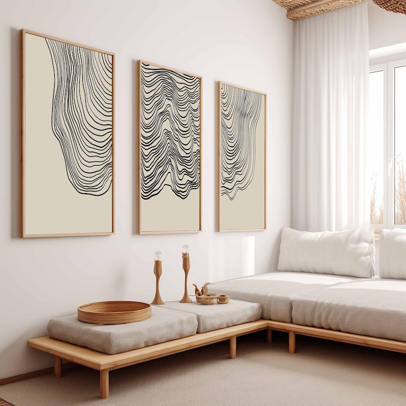 May include: Three framed abstract line art prints with black lines on a cream background. The prints are hung on a white wall above a light brown wooden bench with a white cushion.