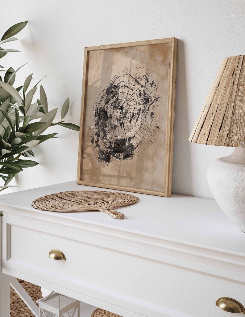 May include: A framed print of a black and white tree ring design on a beige background. The print is hanging on a white wall above a white dresser with a woven fan and a lamp with a woven shade.
