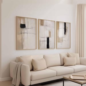 May include: Three framed abstract art pieces. Each features a beige background with black and tan geometric shapes and lines. The frames are a light wood color. The artwork hangs above a cream-colored sofa with matching pillows.