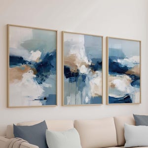 May include: Three framed abstract art prints. Each print features a blend of blue, white, and tan brushstrokes. The artwork is displayed in light-colored wooden frames, creating a cohesive set. The paintings are mounted above a beige sofa.