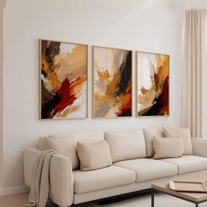 May include: Three framed abstract art pieces with warm tones of brown, orange, and red. The artwork features brushstroke-style designs. The frames are a light wood color, and the art is displayed above a cream-colored sofa.