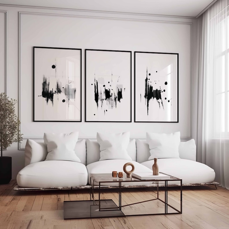 May include: Three black and white abstract art prints in black frames hanging on a white wall above a white couch with white pillows. The couch is in front of a coffee table with a wooden top and a black metal frame.
