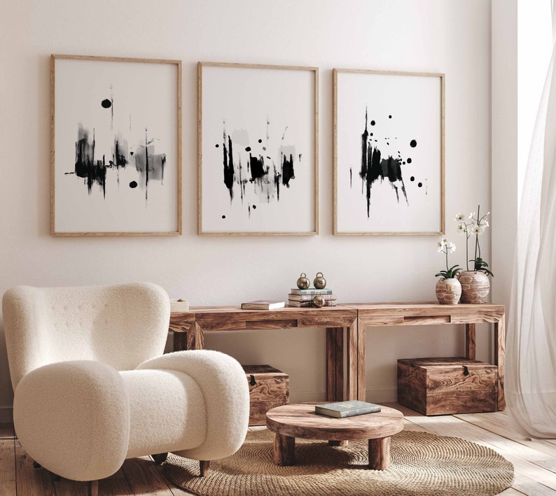May include: Three black and white abstract art prints with wooden frames are displayed on a white wall above a wooden console table with two white ceramic vases and a white armchair.