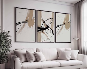 May include: Three framed abstract art prints. Each print features a cream-colored background with black and tan brushstrokes. The frames are black, and the artwork is displayed above a white sofa with matching pillows. A potted plant and a floor lamp are also visible.