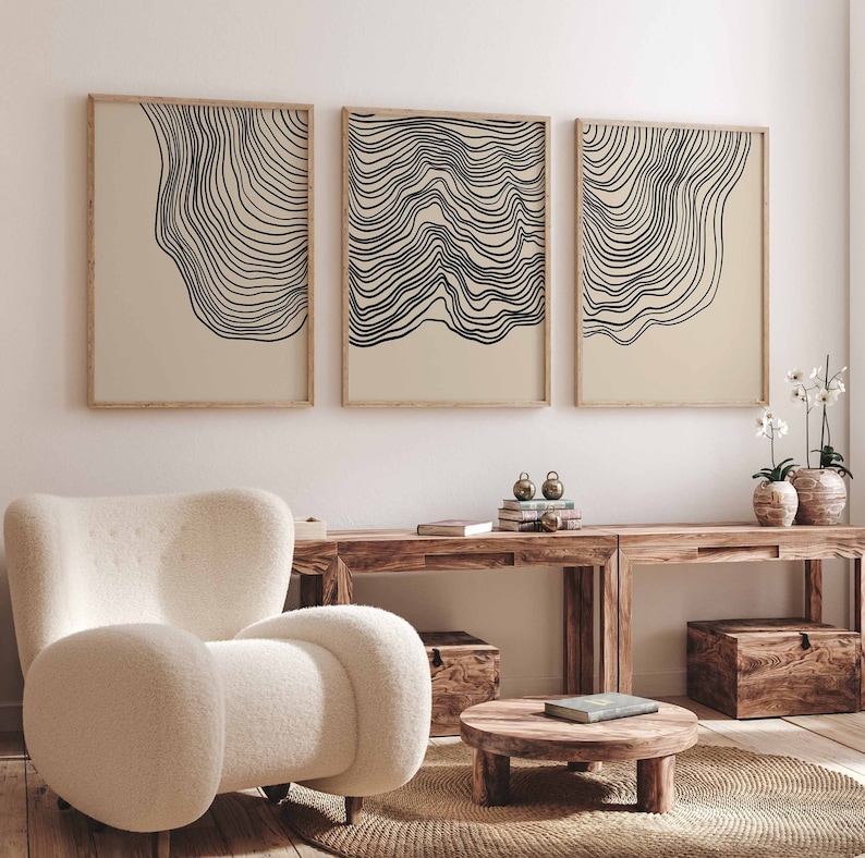 May include: Three abstract line art prints in black and white on a beige background. The prints are framed in natural wood and hung on a white wall above a wooden table and a white armchair.