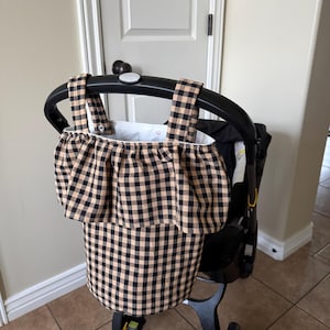 May include: A black and tan gingham patterned stroller organizer bag attached to a black stroller. The bag has a ruffled top and two straps. The stroller has black wheels and a black handle.