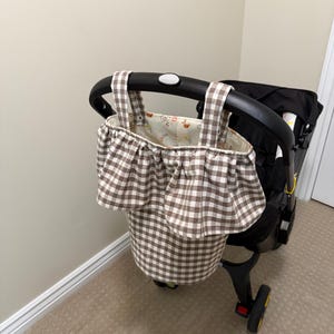 May include: A brown and white gingham fabric storage bag hanging from a black stroller handle. The bag features a ruffled top edge and two fabric straps. The interior lining is a cream-colored fabric with a small, repeating bear and fruit print.