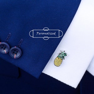 Puede incluir: Un gemelo de piña de plata con hojas verdes y cuerpo amarillo. El gemelo está sobre una camisa blanca con una chaqueta de traje azul.
