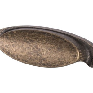 May include: An antique brass cabinet pull with an oval shape and a slightly curved design. The pull has a distressed finish and two visible screws.