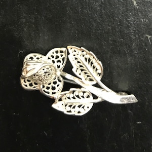 May include: A silver filigree rose brooch with intricate details. The rose is depicted with a single stem and leaves.