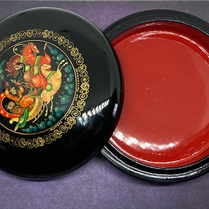 Op de afbeelding: Een ronde zwarte gelakte doos met een rood interieur. Het deksel heeft een kleurrijk geschilderd tafereel van een door paarden getrokken koets met figuren in traditionele Russische volkskledij.