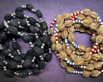 May include: Two necklaces. One is black with silver beads, the other is tan with red, white, and blue beads. The beads are oval-shaped and strung together. The background is a solid purple color.