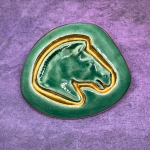 Puede incluir: Un adorno de cerámica verde con un contorno dorado de una cabeza de caballo.