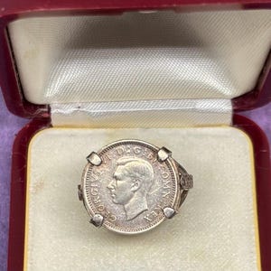 May include: A silver ring featuring a coin with a profile of a man and the text "GEORGIVS VI D:G: BR: OMN: REX". The ring is set in a decorative silver band and is displayed in a red jewelry box.