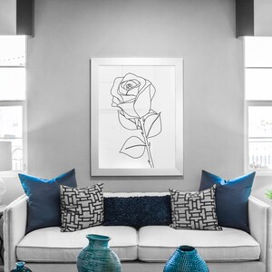 May include: A white sofa with blue cushions in a living room setting. The sofa is in front of a window with white blinds. There is a black and white line drawing of a rose in a white frame on the wall behind the sofa.