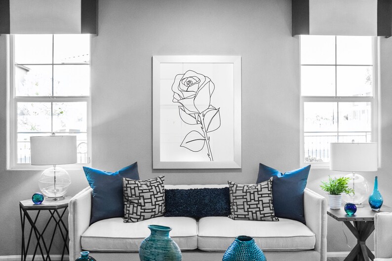 May include: A white sofa with blue cushions in a living room setting. The sofa is in front of a window with white blinds. There is a black and white line drawing of a rose in a white frame on the wall behind the sofa.