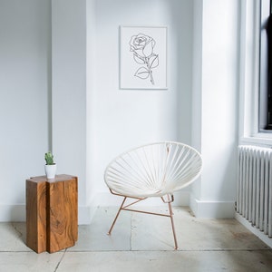 May include: A white chair with a woven design sits in a white room with a wooden side table and a framed line drawing of a rose on the wall.