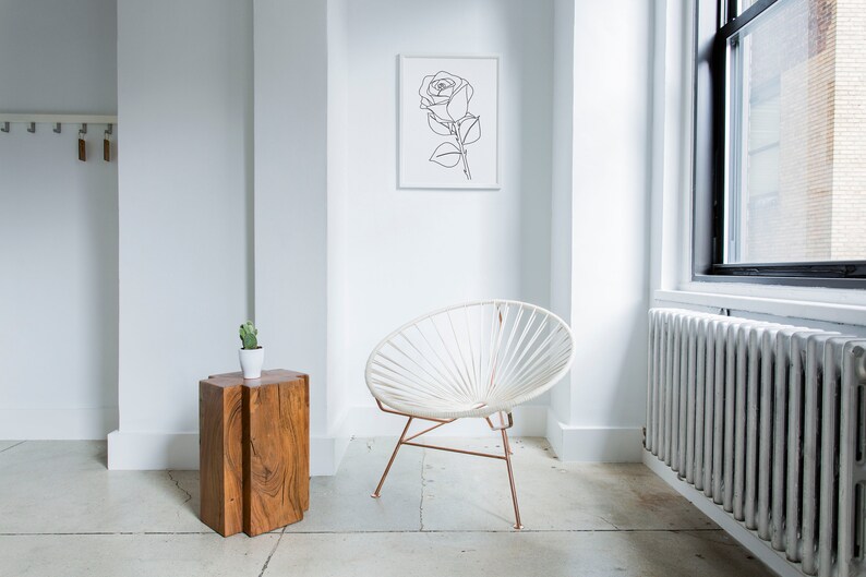 May include: A white chair with a woven design sits in a white room with a wooden side table and a framed line drawing of a rose on the wall.