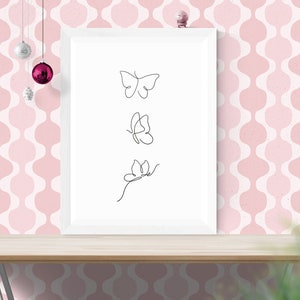May include: A white framed print with three black line drawings of butterflies on a white background. The print is sitting on a wooden table in front of a pink and white patterned wall.