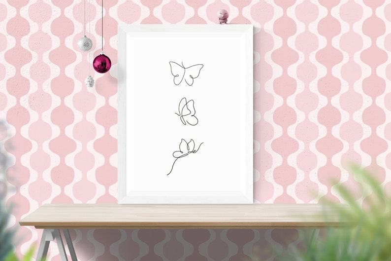 May include: A white framed print with three black line drawings of butterflies on a white background. The print is sitting on a wooden table in front of a pink and white patterned wall.