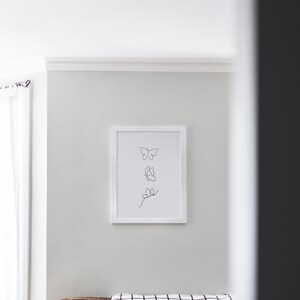 May include: A white framed print with a black line drawing of three butterflies on a white background. The print is hanging on a white wall above a brown couch with a black and white checkered throw blanket and two pillows.