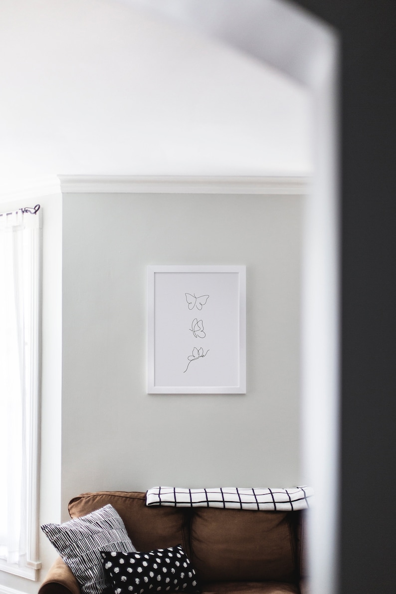 May include: A white framed print with a black line drawing of three butterflies on a white background. The print is hanging on a white wall above a brown couch with a black and white checkered throw blanket and two pillows.