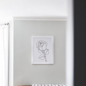 May include: A white framed print of a black line drawing of a single rose with leaves and a stem. The print is hanging on a white wall above a brown sofa with a black and white checked throw blanket and black and white patterned cushions.