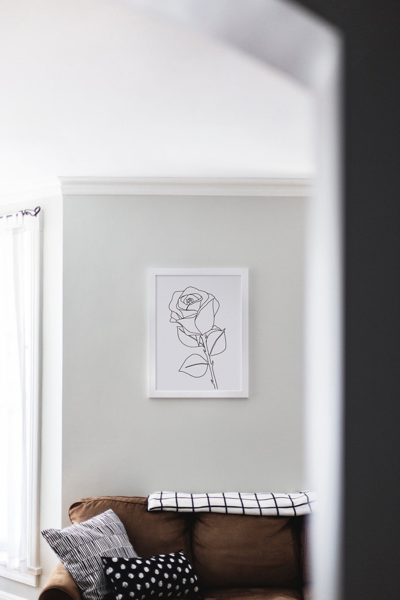 May include: A white framed print of a black line drawing of a single rose with leaves and a stem. The print is hanging on a white wall above a brown sofa with a black and white checked throw blanket and black and white patterned cushions.