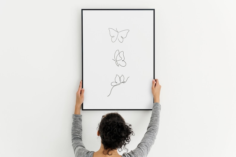 May include: A person hangs a framed print with a black frame on a white wall. The print features three black line drawings of butterflies.