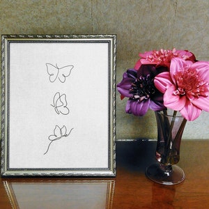 May include: A framed print with three simple line drawings of butterflies. The print is on a white background and is framed in a silver frame. The frame is sitting on a wooden table next to a vase of pink and purple flowers.