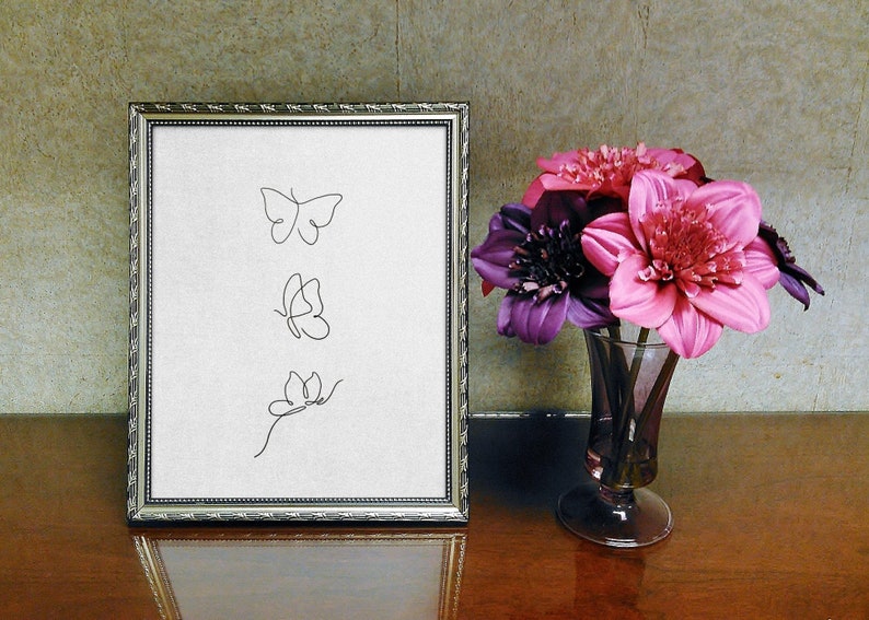 May include: A framed print with three simple line drawings of butterflies. The print is on a white background and is framed in a silver frame. The frame is sitting on a wooden table next to a vase of pink and purple flowers.