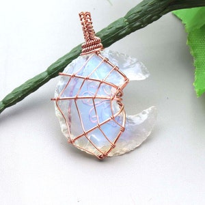 Op de afbeelding: Een koperen draad gewikkeld rond een witte, iriserende maansteen hanger. De hanger is gevormd als een halve maan.