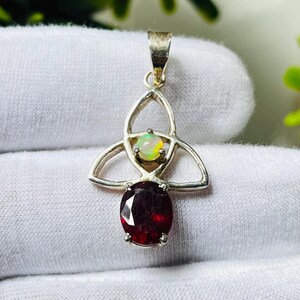 May include: A silver pendant with a red gemstone and a white opal gemstone. The pendant is in the shape of a Celtic knot.