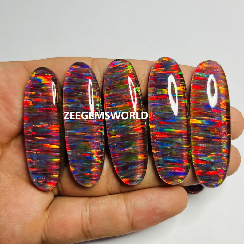 May include: Five oval-shaped, iridescent opal cabochons with a rainbow of colors, including red, orange, yellow, green, blue, and purple. The cabochons are set in a clear, glassy material.  ZEEGEMSWORLD is printed on the image.