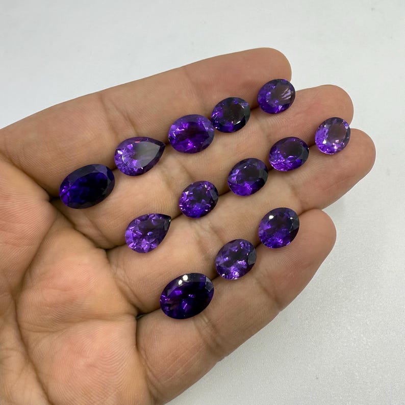 May include: A collection of polished, faceted gemstones in various shapes, including oval and pear cuts. The gems display a deep purple hue with internal light reflections. The stones are held in a hand against a white background.