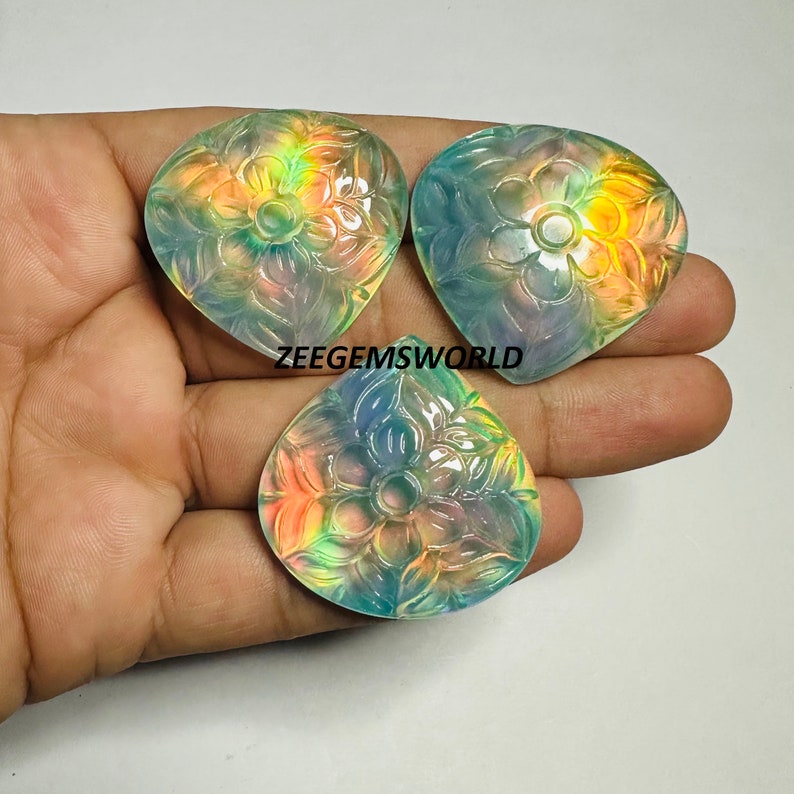 May include: Three iridescent, carved gemstone pendants in the shape of a teardrop with a floral design. The gemstones are a pale blue-green color with rainbow iridescence.