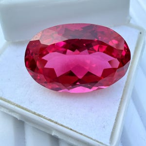 May include: A faceted, oval-shaped gemstone with a deep pink hue. The gem is set against a white background, showcasing its clarity and brilliance. The cut of the stone creates multiple facets, reflecting light and enhancing its color.