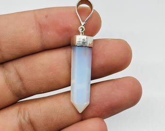 Puede incluir: Un colgante de cristal azul claro con una fianza plateada. El cristal tiene forma hexagonal alargada con una punta. El colgante está sujeto por una mano sobre un fondo blanco.