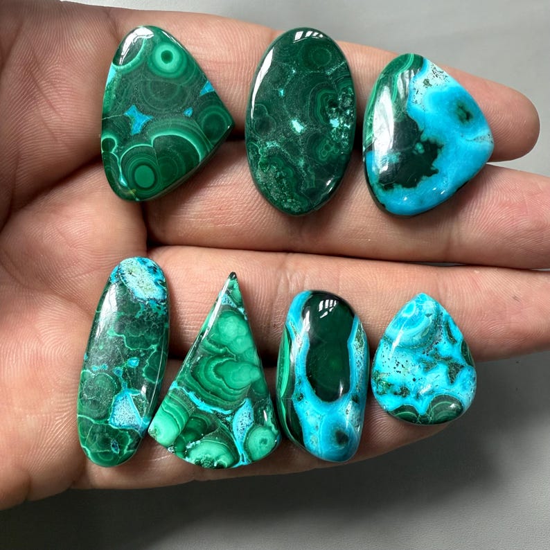 May include: A collection of polished gemstones in various shapes, including oval, teardrop, and triangular. The stones display a mix of deep green and turquoise blue colors with unique swirling patterns. The gemstones are likely used for jewelry or decorative purposes.