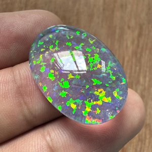 Könnte beinhalten: Ein ovaler Opal-Edelstein mit einem durchscheinenden, irisierenden Aussehen. Der Stein zeigt ein Farbspiel mit grünen, gelben und blauen Blitzen. Der Opal wird in einer Hand gehalten und zeigt seine polierte Oberfläche und die inneren Muster.