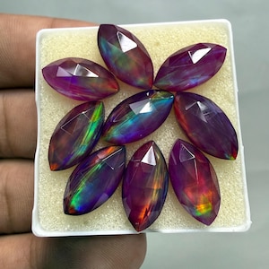 May include: Eight marquise-cut gemstones arranged in a white display box. Each stone features a deep purple base color with iridescent flashes of green, blue, and orange light reflecting across the faceted surfaces in a vibrant, multi-colored display.