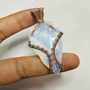 May include: A copper wire wrapped pendant featuring a white opal stone. The opal is a rough, irregular shape and has a milky white color. The copper wire is wrapped around the opal in a delicate and intricate design.